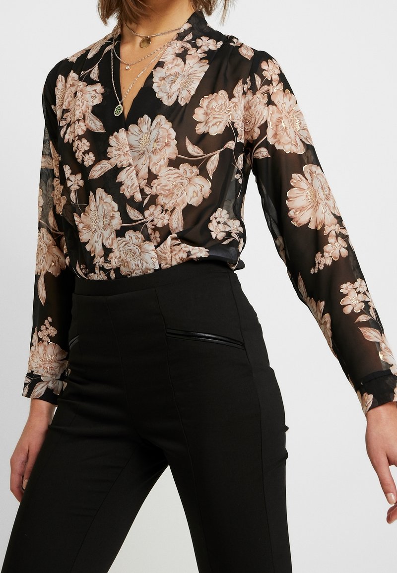 Czarna kwiecista bluzka wykonana z przezroczystej tkaniny z motywami różowych i kremowych kwiatów, uzupełniona długimi rękawami i dekoltem w kształcie litery V, zestawiona z czarnymi spodniami.