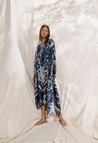 Robe à motifs bleu marine et blancs avec un design fluide, présentant une silhouette ouverte et un ourlet asymétrique. Le mannequin porte des sandales à brides.