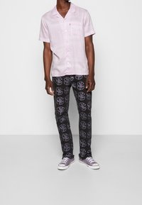 Man wearing light purple patterned short-sleeve shirt, black pants with purple circular designs, and purple low-top sneakers standing against white wall.