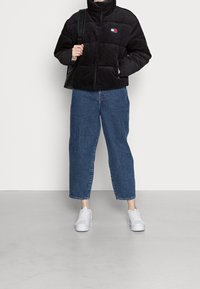 Černá kordurová péřová bunda s vysokým límcem, s logem, v kombinaci s modrými rovnými džíny a bílými teniskami.
