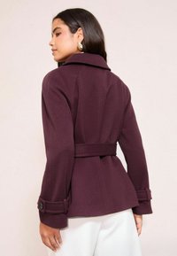 Bordo spalvos siuvinėta švarkas su struktūruotais pečiais, su diržu juosmenyje ir atsegamais rankogaliais. Tekstūruotos medžiagos suteikia dizainui gylio.