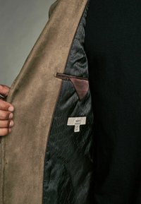 Tissu extérieur en cuir suédé brun avec une doublure noire texturée. Poche intérieure et étiquette visibles, avec un motif subtil.