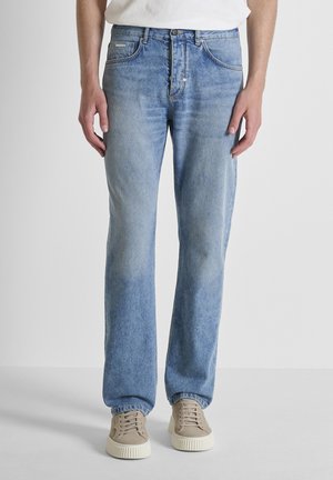 Man die lichtblauwe jeans met rechte pijpen en beige sneakers draagt, staand tegen een effen lichte achtergrond.