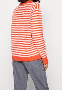 Oranžovo-bílé pruhované svetr s měkkou, chlupatou texturou. Design má zaoblený výstřih a žebrovaný lem. Spárováno se šedými džíny.