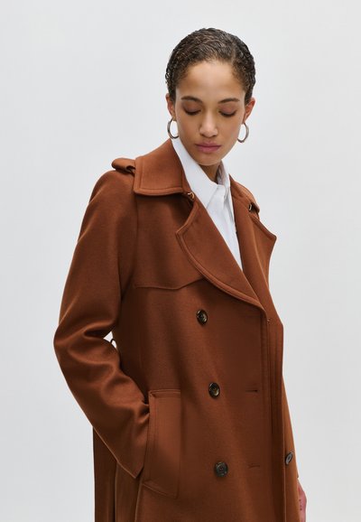 Woman with braided hair wearing gold hoop earrings and a brown double-breasted coat over a white collared shirt, looking down with hand in pocket.