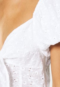 Blusa bianca con maniche a sbuffo caratterizzata da un tessuto testurizzato con ricami e dettagli a occhiello, che presenta una scollatura a V e un design aderente.