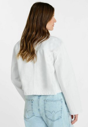 Vrouw met lang bruin haar draagt een witte cropped jas en lichtblauwe spijkerbroek, zijwaarts gezien tegen een effen witte achtergrond.