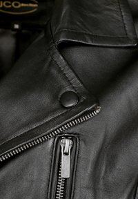 Schwarze Lederjacke mit Reverskragen, ausgestattet mit silbernen Reißverschlüssen und einem Druckknopf. Das Leder hat eine glatte Textur und einen subtilen Glanz.