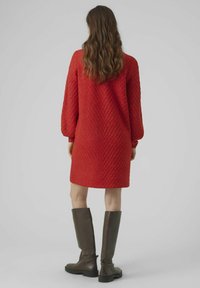 Abito maglione rosso con un motivo a zig zag testurizzato, maniche lunghe e taglio dritto. Indossato con stivali in pelle marrone al ginocchio.