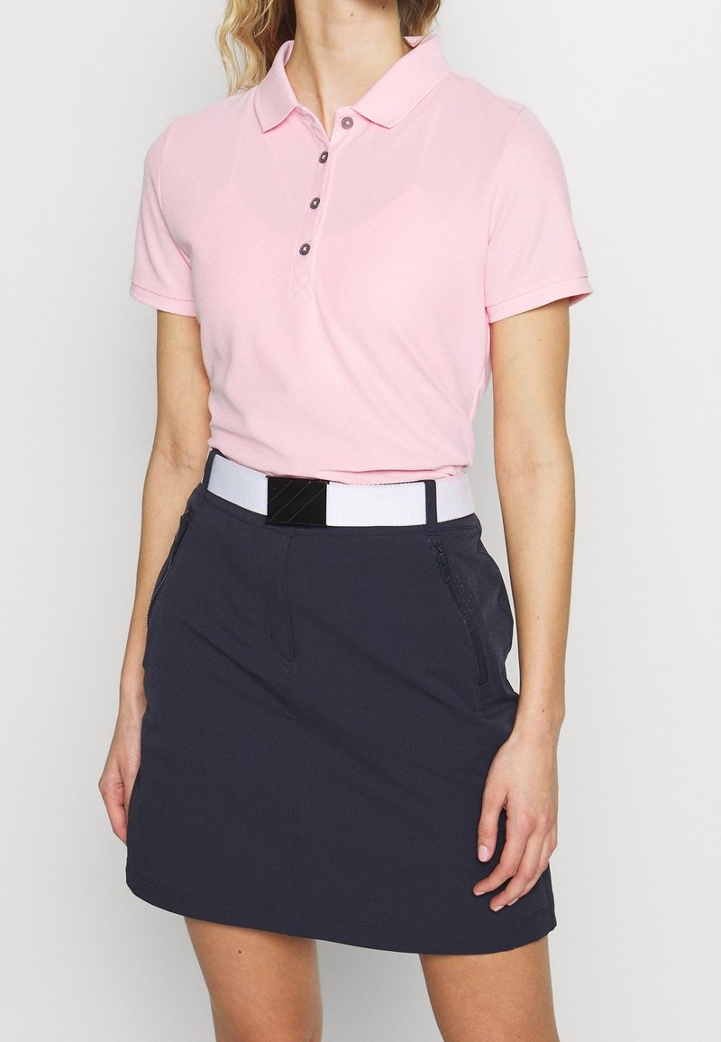 Polo rose clair avec un col et trois boutons, associé à une jupe bleu marine avec des poches zippées et une ceinture blanche.