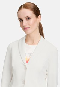 Biały strukturalny żakiet z kołnierzem szalowym, z dwoma guzikami, noszony na jasnym topie z kolorowym wzorem graficznym.