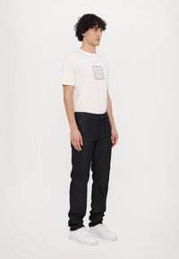 Tricou alb cu gât rotund și logo pe față, purtat cu blugi de denim bleu închis și adidași albi. Design simplu, croială slim, detalii minime.