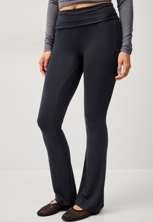 Zwarte flared leggings gemaakt van rekbare stof, met een brede, gevouwen tailleband en een gladde textuur, gecombineerd met zachte zwarte flats.