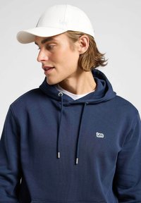 Biała czapka baseballowa z haftowanym logo "Lee", w połączeniu z granatowym dresowym swetrem z kapturem, posiadającym sznurki i srebrne akcenty.