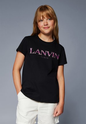 Jong meisje met lang blond haar, gekleed in een zwart Lanvin Paris t-shirt en witte shorts, staande tegen een effen grijze achtergrond.