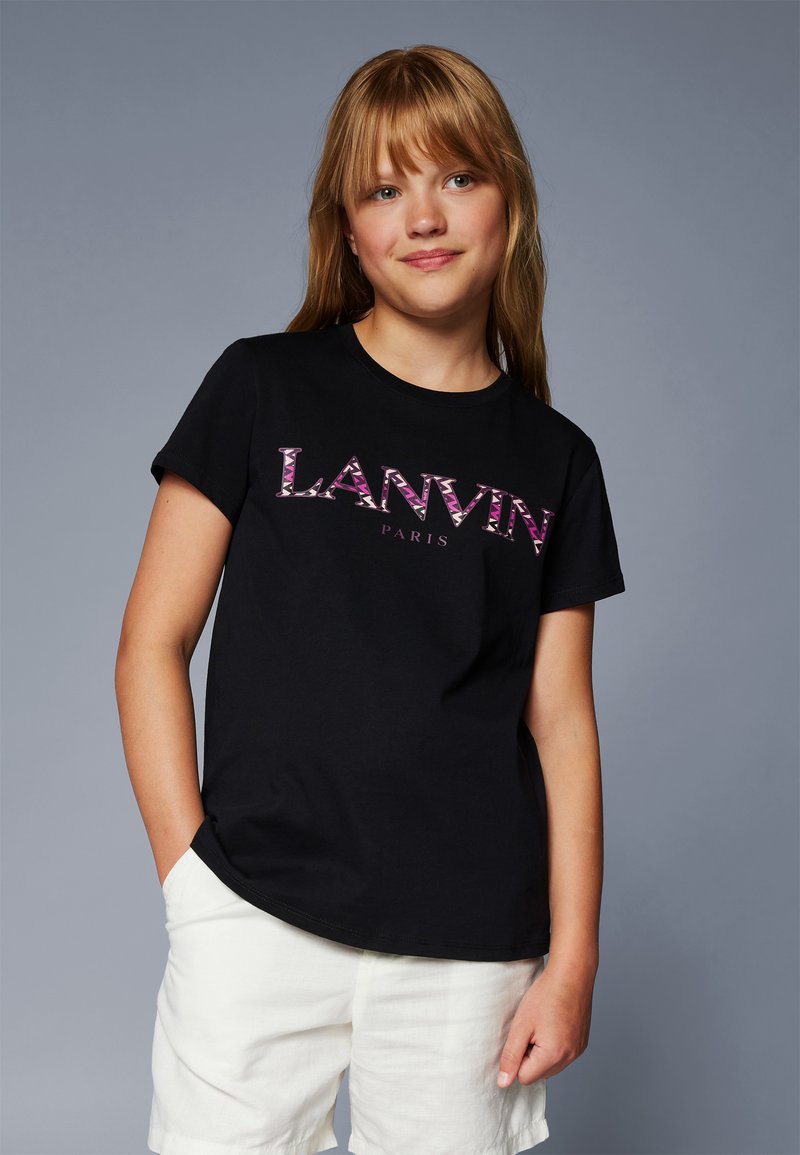 Jeune fille aux longs cheveux blonds portant un t-shirt noir Lanvin Paris et un short blanc, se tenant devant un fond gris uni.