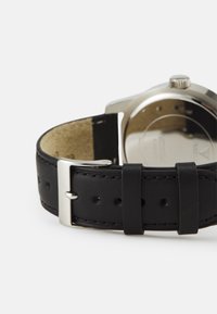 Schwarzes Lederarmband mit silberner Metallschließe, mit Nähdetails und Löchern zur Anpassung, das Gehäuse der Uhr teilweise sichtbar.
