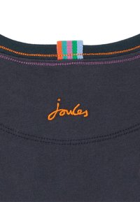 Tela azul marino con bordado naranja "joules" debajo de un lazo de tela a rayas multicolor y costuras naranjas y moradas en la línea del cuello.