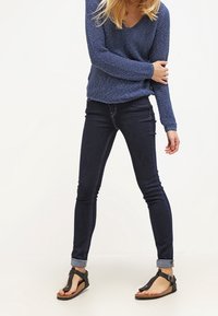 Person trägt einen blauen Strickpullover, dunkle enge Jeans mit Umschlag und schwarze Zehensandalen, steht mit einem Arm quer über dem Körper.