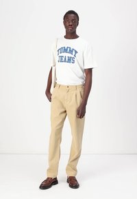 Hvit t-skjorte med blå "TOMMY JEANS" tekst, beige bukser, brune sko, og en skulderveske, stående på en hvit bakgrunn.