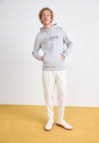 Lichtgrijze hoodie met een gedrukt zwart logo, een voorzak en een trekkoord. Gecombineerd met witte tapse broek met ritszakken en witte sneakers.