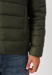 Olivengrønn puffjakke med horisontal quilted design, en teksturert overflate og en utvendig lomme, båret over denim.