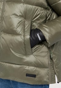 Olivgrön vadderad jacka med blixtlåsförslutning, sidofickor och texturerad yta. Inkluderar en svart läderdetalj på ärmen.