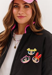 Čierna prešívaná bunda s ružovým lemovaním, s tromi vyšívanými nášivkami s kreslenými postavičkami na hrudi. Pod ňou biela košeľa.