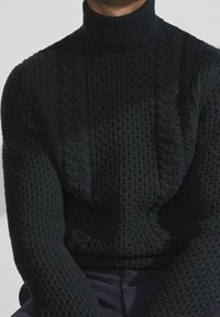 Ciemnozielony, sweter z golfem, wykonany na drutach z wzorem warkocza i prążkowanym kołnierzem. Krój jest luźny, podkreślając grubość materiału.