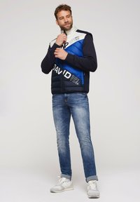 Gilet imbottito con pannelli blu, navy e bianchi, dotato di un colletto alto e chiusura con zip. Abbinato a jeans blu e scarpe da ginnastica chiare.