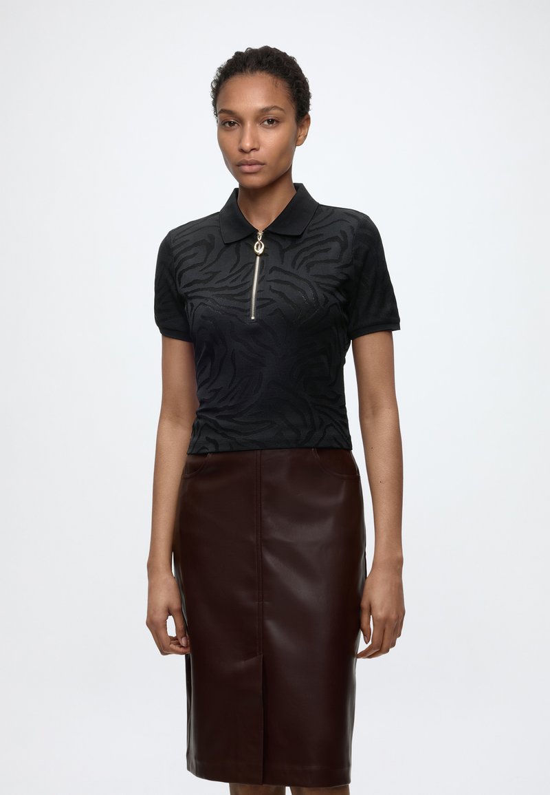 Γυναίκα που φοράει μαύρο κοντομάνικο πόλο με διακριτικό ζέβρα μοτίβο και χρυστό φερμουάρ, συνδυασμένο με σκούρα καφέ δερμάτινη φούστα μήκους μέχρι το γόνατο.