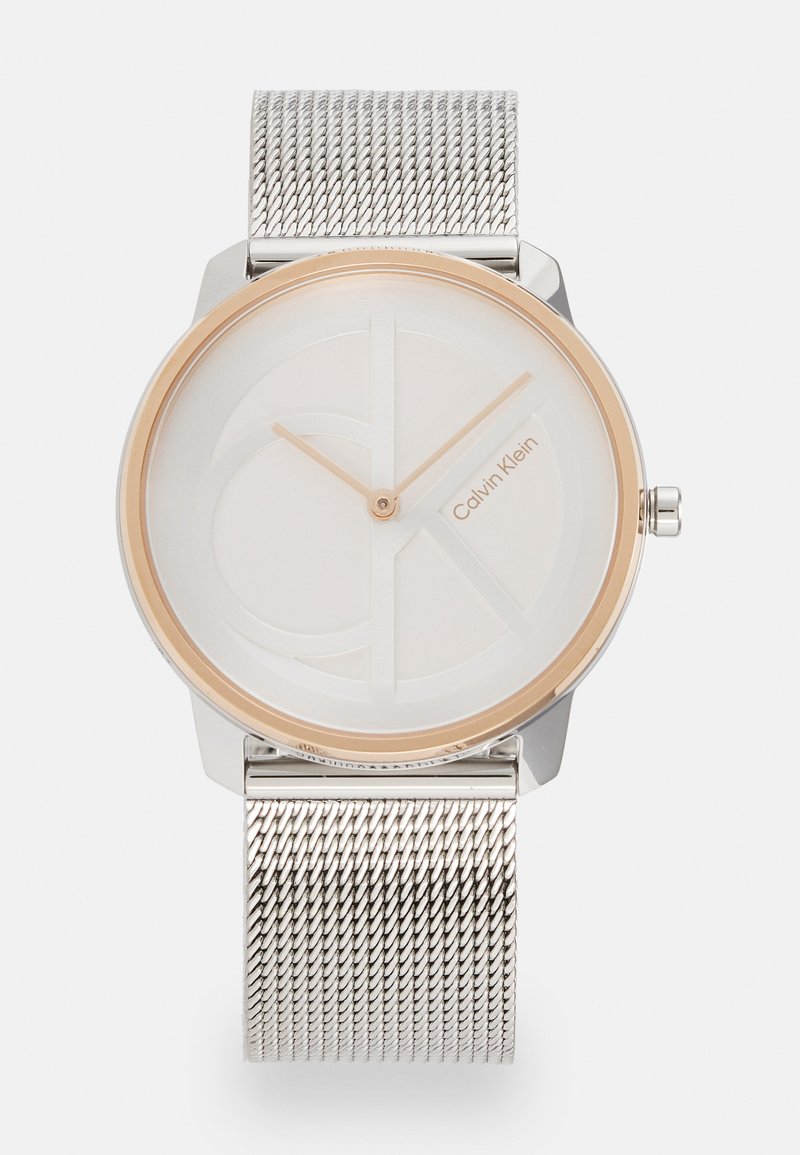 Montre ronde en argent avec un cadran en or, face blanche minimaliste embossée du logo, et bracelet en maille argentée. Aiguilles des heures et des minutes simples.