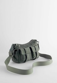 Sac bandoulière vert avec deux poches avant à boucles et une sangle large réglable sur un fond clair uni.