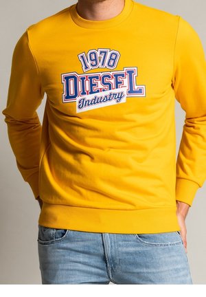 Sweat-shirt jaune avec le texte "1978 Diesel Industry" porté avec un jean bleu clair, mains dans les poches, fond neutre.
