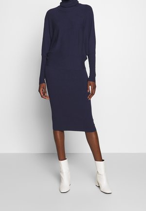 Femme portant une robe à col roulé bleu marine à manches longues avec une jupe longueur genou et des bottines blanches, debout sur un fond uni.