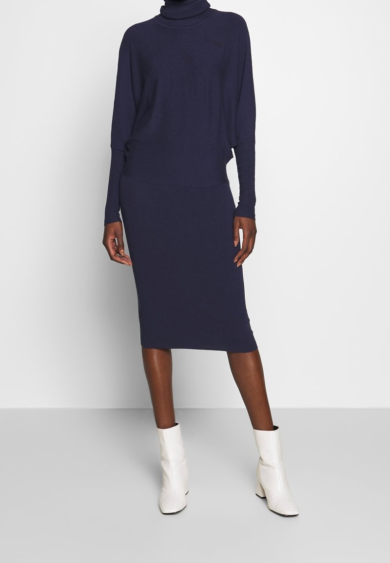 Femme portant une robe à col roulé bleu marine à manches longues avec une jupe longueur genou et des bottines blanches, debout sur un fond uni.