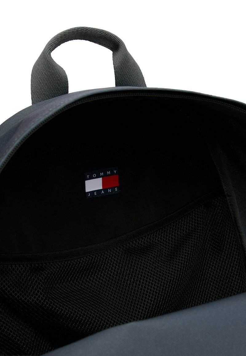 Black backpack interior featuring a mesh pocket and a tag with the "TOMMY JEANS" logo in red, white, and blue on a dark background.