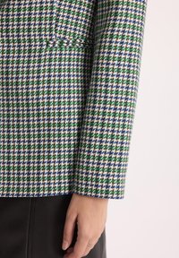 Rękaw kurtki w wzór houndstooth w kolorach zielonym, niebieskim i białym, o teksturowanym wykończeniu; posiada dopasowany krój i wyraźne detale szwów.