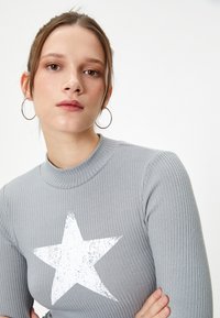 Pull gris à côtes avec un imprimé étoile usé blanc sur le devant et un col haut. Mannequin portant des boucles d'oreilles argentées en forme de créoles.