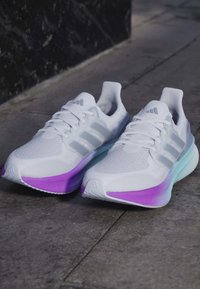 Λευκά παπούτσια τρεξίματος Adidas με μοβ και μπλε διαβαθμισμένες σόλες τοποθετημένα σε πέτρινο δάπεδο.
