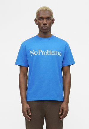Mand med kort blond hår iført blå T-shirt med hvid tekst "No Problemo" og brune bukser, stående foran en ensfarvet hvid baggrund.