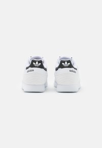 Baltās Adidas kedas ar melnu papēdi un logo, ar teksturētu augšpusi, noapaļotu formu un gludu zoli.