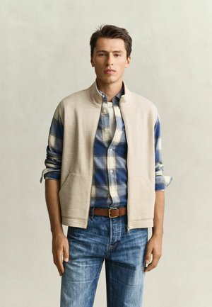 Giovane uomo in piedi di fronte a noi, indossa un gilet beige con zip sopra una camicia a quadretti blu e bianca con le maniche risvoltate e jeans blu.