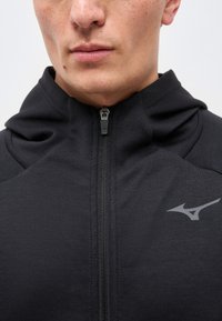 Κοντινό πλάνο του κάτω μέρους του προσώπου και του λαιμού που φοράει μια μαύρη φούτερ με φερμουάρ και ένα μικρό γκρι λογότυπο στο στήθος.