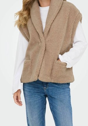 Persoon draagt een beige zachte mouwloze vest over een wit shirt met lange mouwen, met de handen gedeeltelijk in de zakken van de vest en blauwe spijkerbroek.
