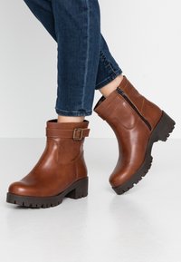 Botas de tobillo de cuero marrón con cremallera lateral, acento de hebilla y suela de goma gruesa. Textura suave y diseño de punta redondeada.