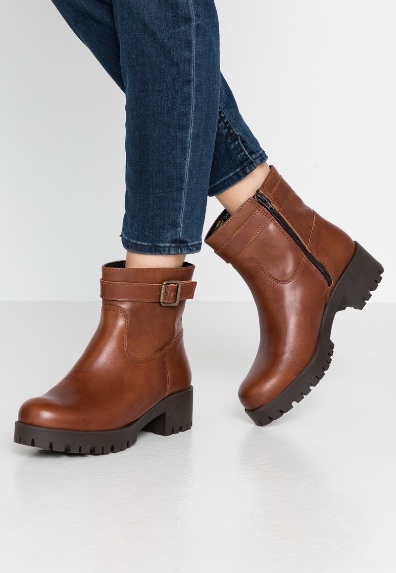 Botas de tobillo de cuero marrón con cremallera lateral, acento de hebilla y suela de goma gruesa. Textura suave y diseño de punta redondeada.