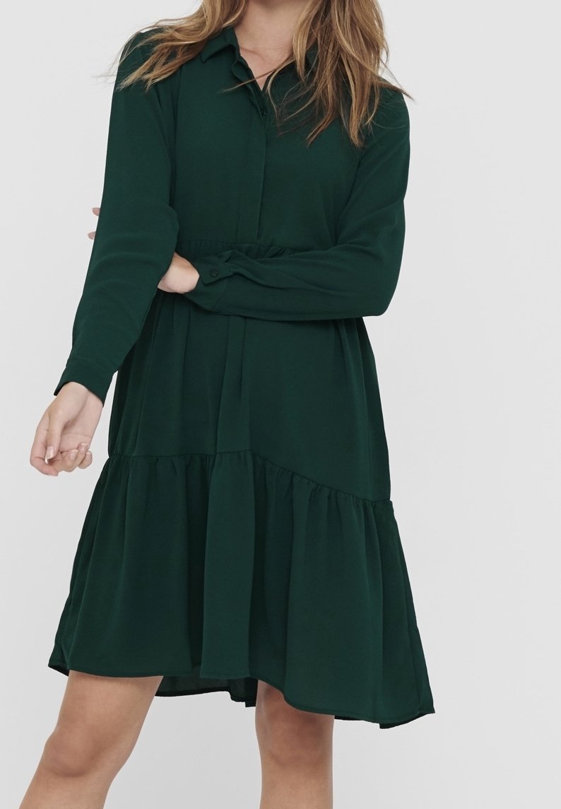 Femme portant une robe verte foncée à manches longues avec une jupe à volants et un col, debout devant un fond clair uni.