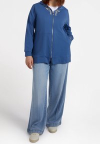 Blue zip-up hoodie with a hood, featuring decorative embellishments along the neckline. Paired with wide-leg light blue denim jeans.