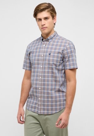 Junger Mann trägt ein kurzärmliges Hemd mit blau-orange-weißem Karomuster und steht vor einem schlichten hellen Hintergrund.